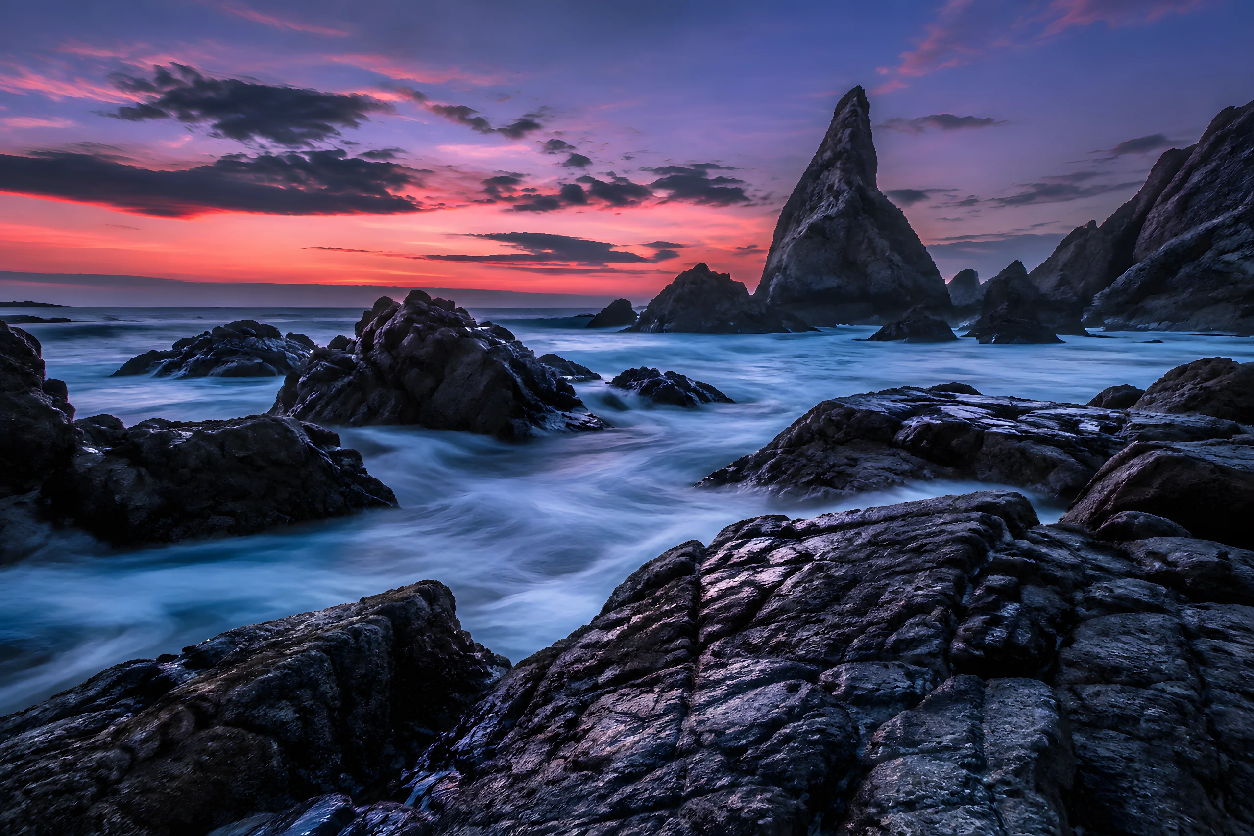 Long exposure of a rocky coast at blue hour, the sea blurred to mist around dark rocks and a pointed sea stack under a pink and violet sky. Sample image, AI-generated.