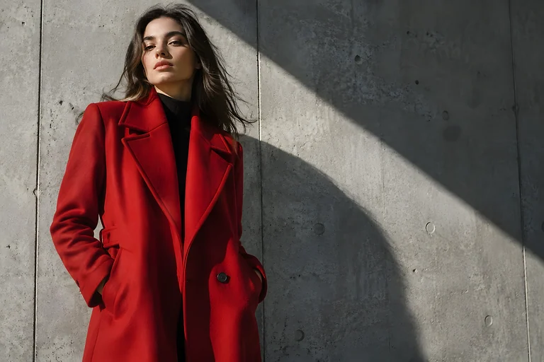 A woman in a bold red coat leaning against raw concrete, with a hard diagonal shadow across the wall. Sample image, AI-generated.