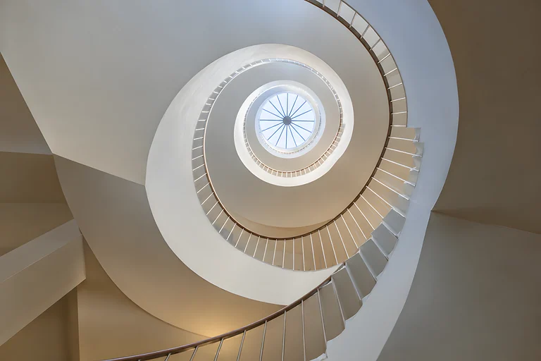 A white spiral staircase photographed from below, curving up towards a round skylight. Sample image, AI-generated.